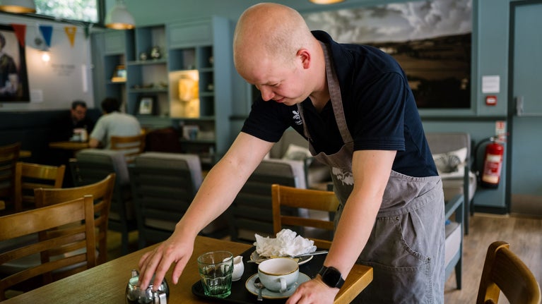 Young volunteer clearing a tray from a table in the cafe at Sizergh. He is wearing a grey apron with the white National Trust oak leaf logo on.